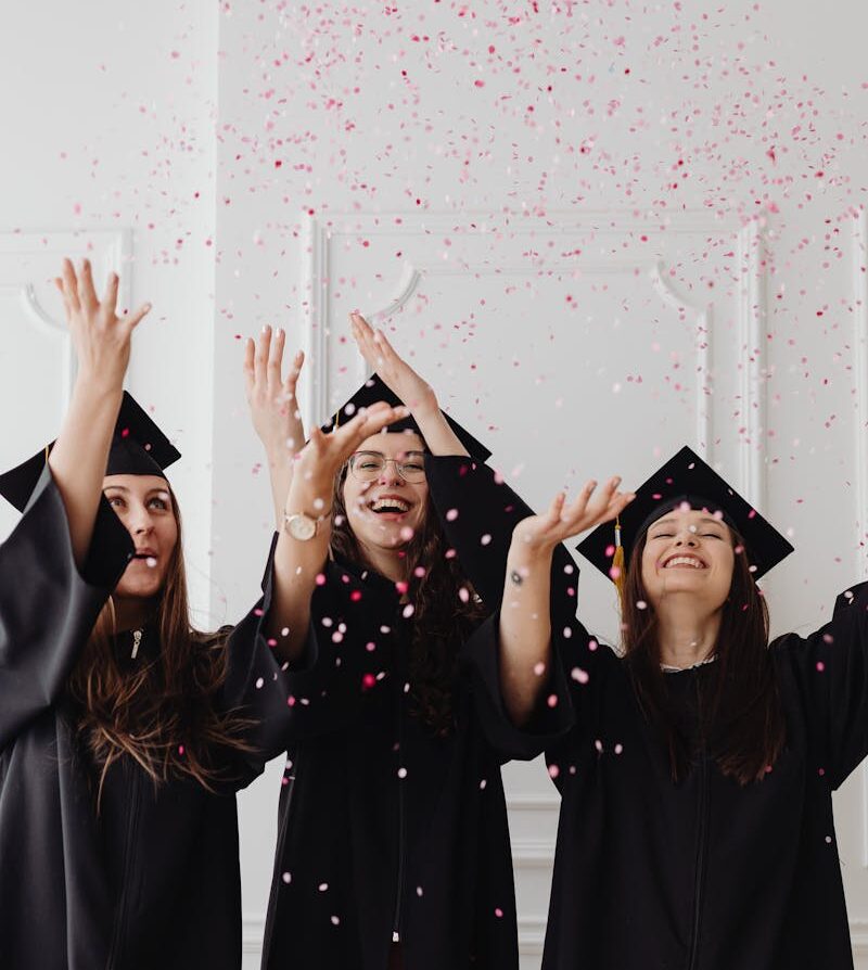 Three young women celebrating graduation by throwing confetti indoors.